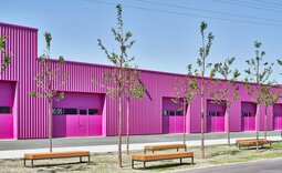 Hallenfassade in kräftigem Verkehrspurpur mit nummerierten Toren, vorgelagerten Bäumen und Sitzbänken entlang der Verkehrsfläche